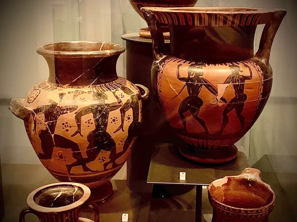 Ancient Greek-style ceramic vases with painted mythological scenes displayed in Chiusi museum, associated with Etruscan trade and burial culture.